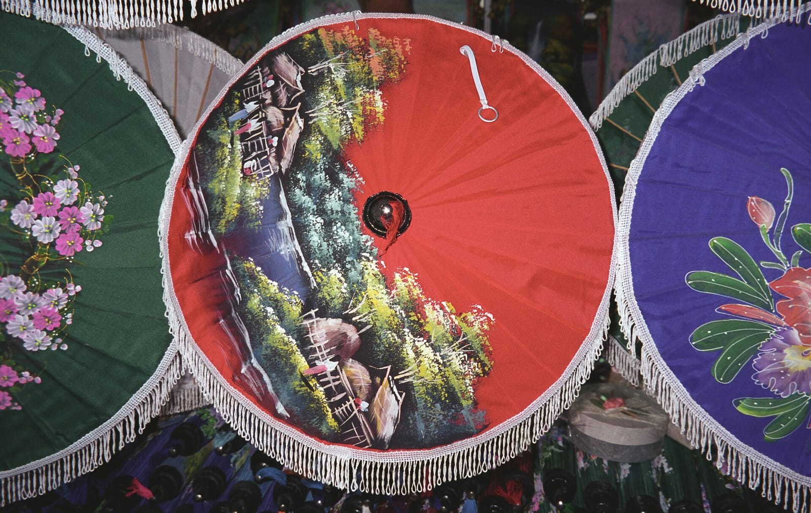 Hand-painted Bo Sang rice-paper umbrellas with white fringes displayed side by side, one showing a river landscape motif