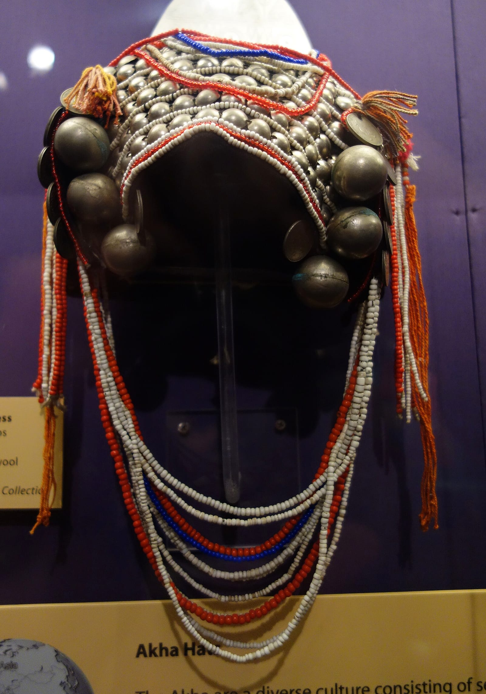 Akha married woman's headdress heavy with silver bosses, coins and beaded tassels, displayed in a museum case