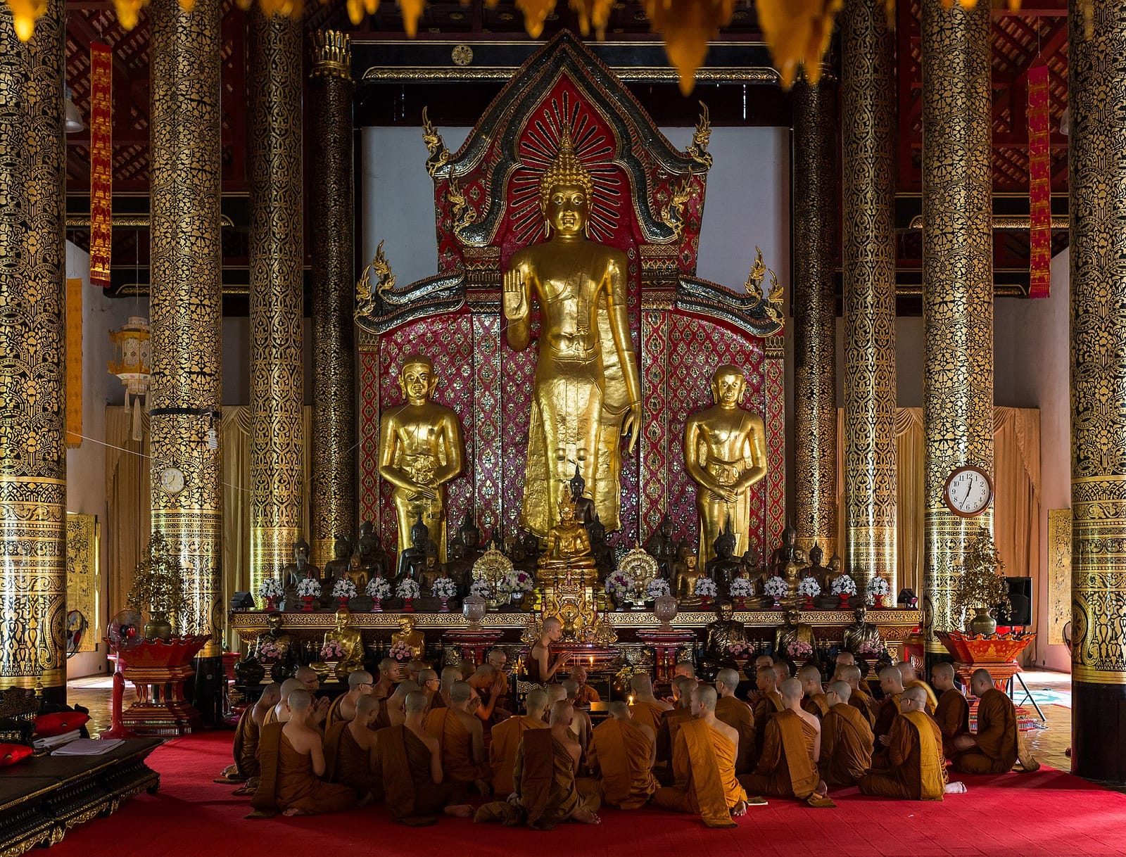 The large standing Buddha image inside the principal viharn, attended by smaller seated figures, with monks gathered before the altar