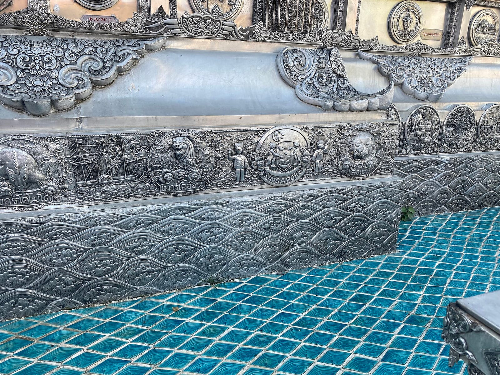 Embossed medallion panels along the lower outer wall of the silver ubosot, showing animal and national emblems above the wave-pattern base