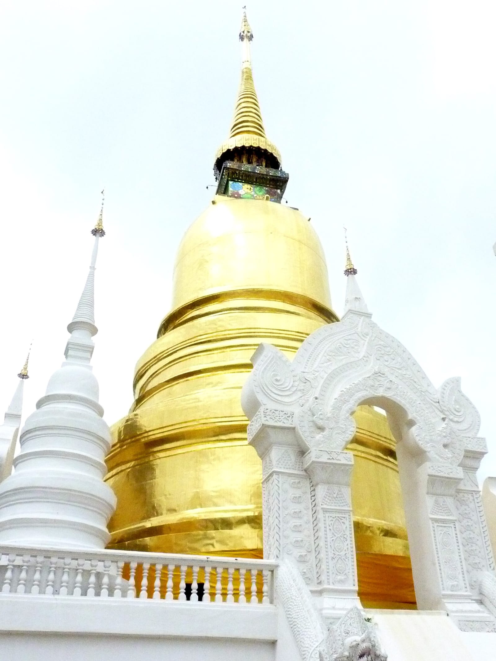 The gilded principal chedi of Wat Suan Dok rising behind a white memorial arch and surrounding white chedis