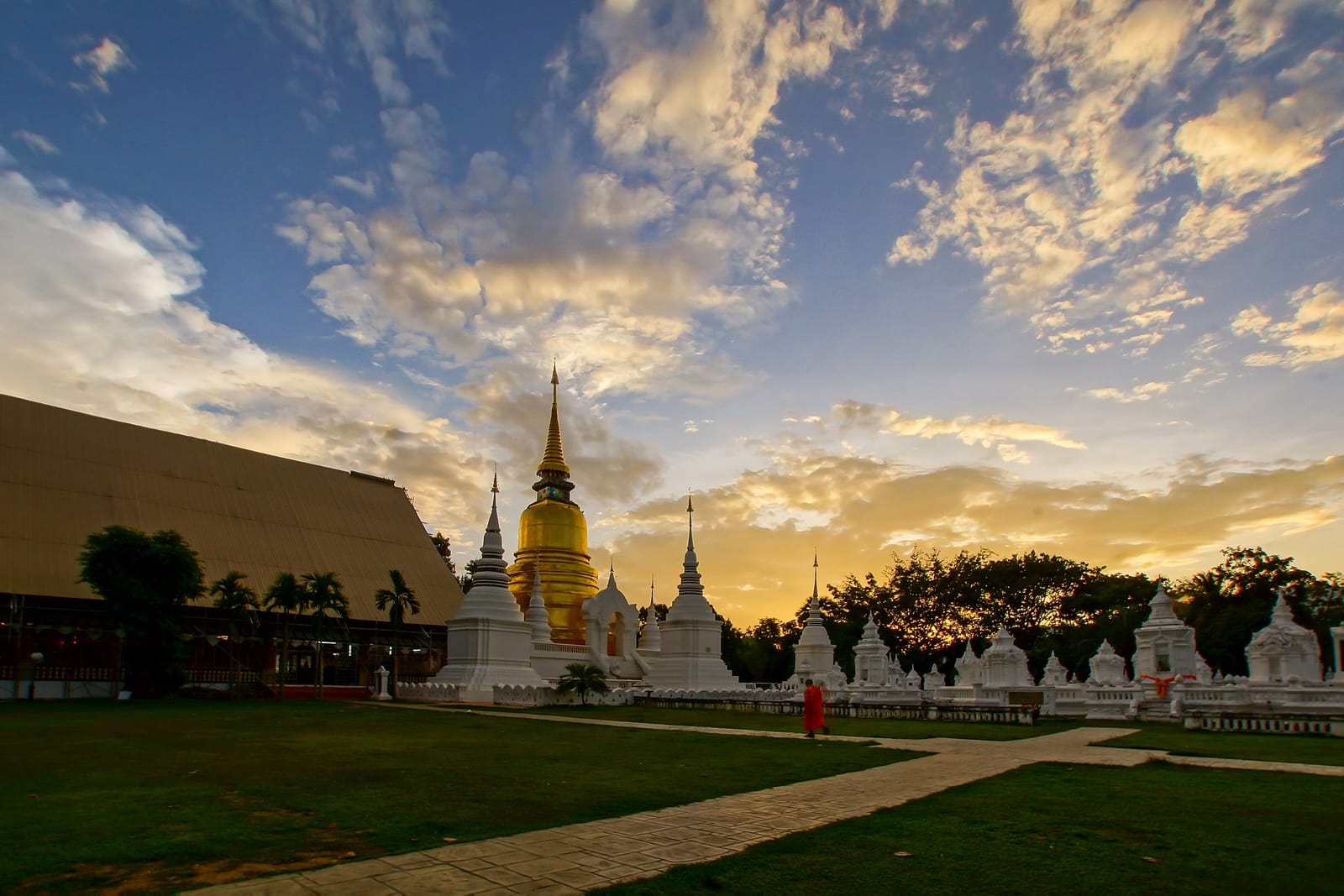 The gilded chedi and white royal memorials of Wat Suan Dok against the evening sky at sunset, with a monk crossing the lawn