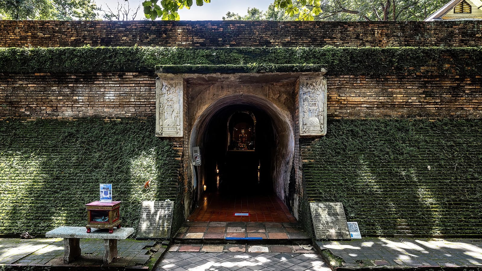Arched brick entrance to the underground tunnels at Wat Umong, moss-covered, with the lit Buddha shrine visible inside